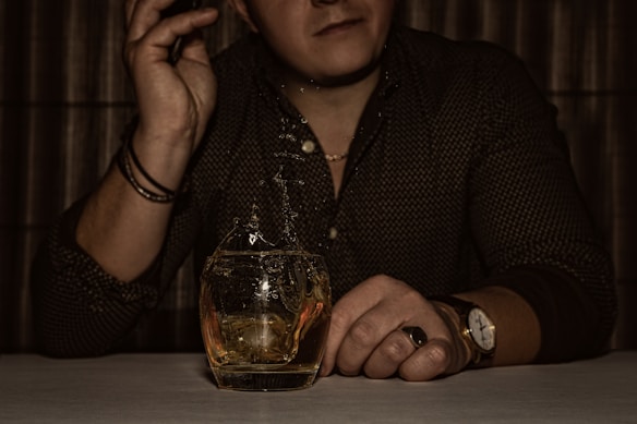 A person seated at a table holding a small glass of whiskey with a splash captured mid-air. The person's head is partially visible, and they are wearing a patterned shirt and jewelry, including bracelets and a watch.