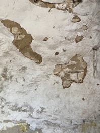 A weathered wall with patches of exposed brick and plaster. The surface is uneven with several areas showing signs of cracking and peeling paint. The texture is rough and the underlying structure is partially visible.
