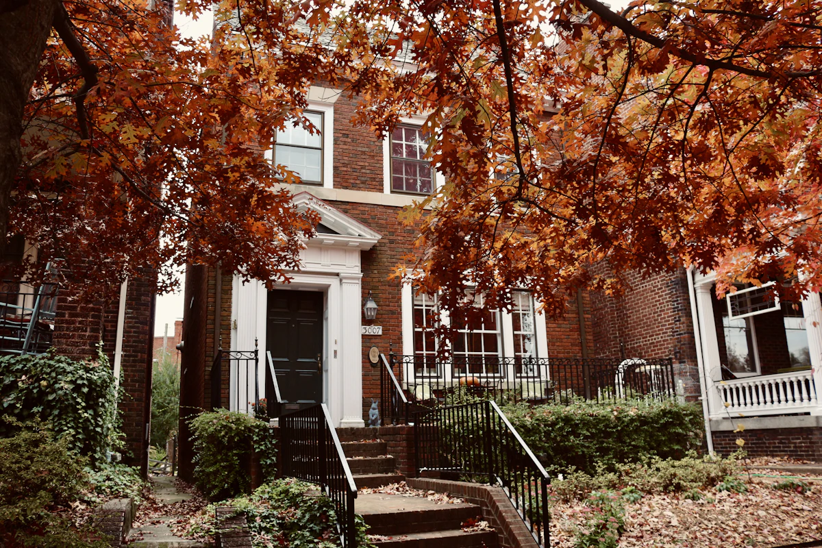 A red-brick house with a white porch behind maple trees in autumn colour