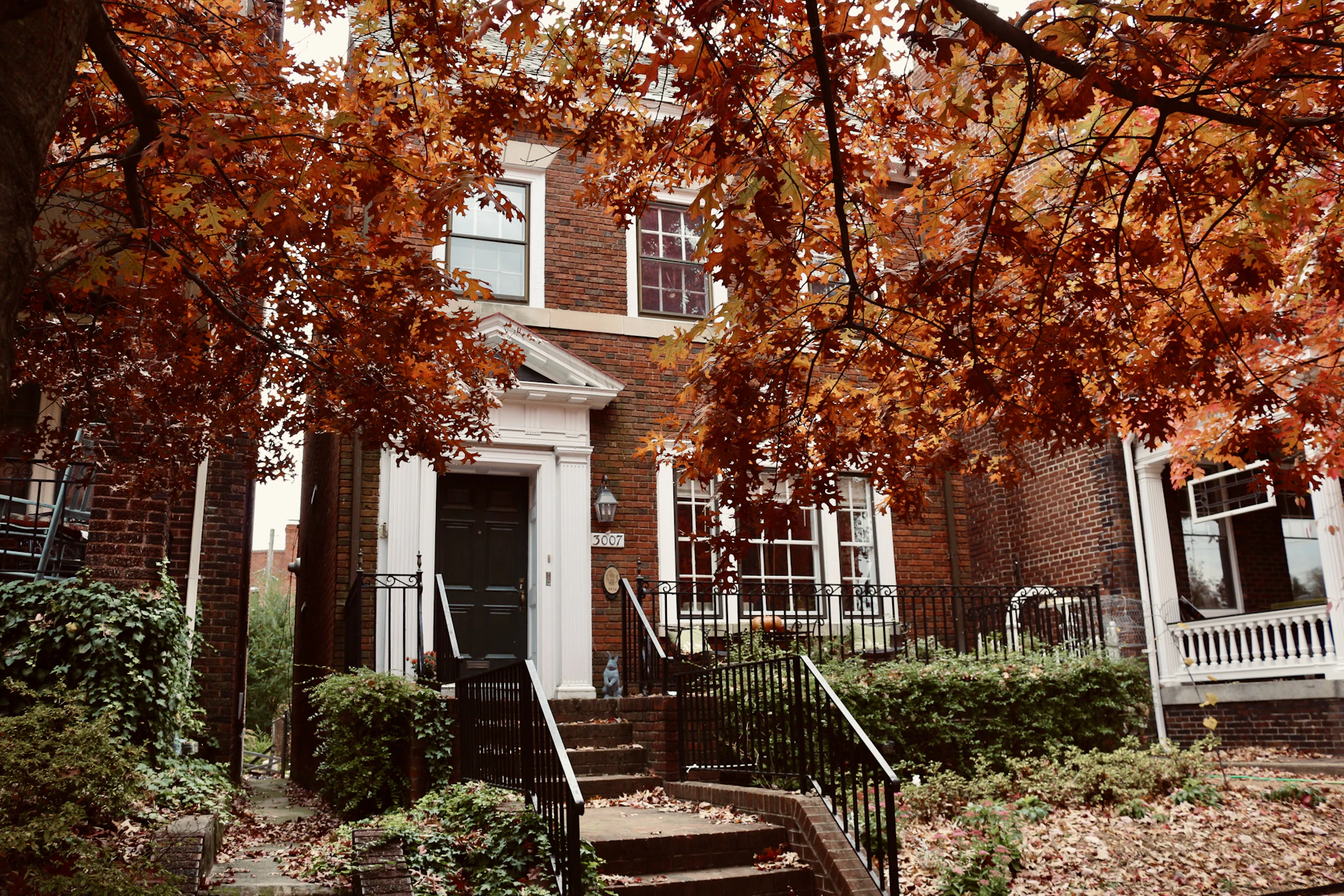 A red-brick house with a white porch behind maple trees in autumn colour