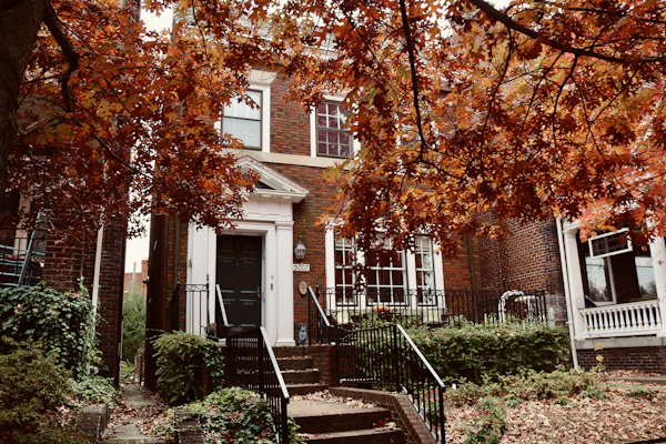 A red-brick house with a white porch behind maple trees in autumn