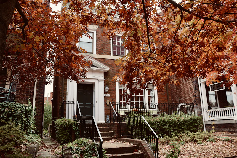 A red-brick house with a white porch behind maple trees in autumn colour