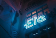 A dimly lit urban scene featuring a neon sign with the letters 'Eic'. The setting has a moody, artistic vibe with a mural on the wall depicting abstract shapes and lines. Several windows reflect some of the neon light, adding depth to the scene.