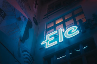 A dimly lit urban scene featuring a neon sign with the letters 'Eic'. The setting has a moody, artistic vibe with a mural on the wall depicting abstract shapes and lines. Several windows reflect some of the neon light, adding depth to the scene.