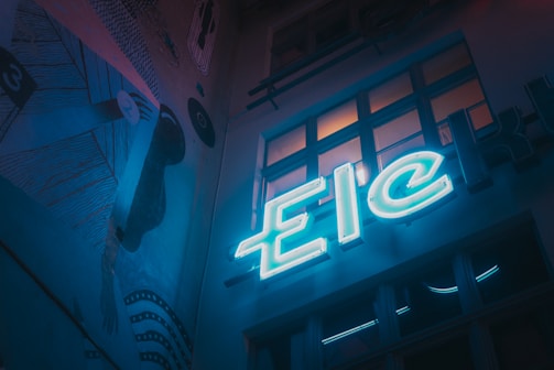 A dimly lit urban scene featuring a neon sign with the letters 'Eic'. The setting has a moody, artistic vibe with a mural on the wall depicting abstract shapes and lines. Several windows reflect some of the neon light, adding depth to the scene.