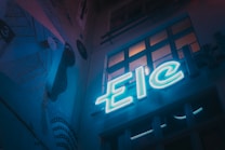 A dimly lit urban scene featuring a neon sign with the letters 'Eic'. The setting has a moody, artistic vibe with a mural on the wall depicting abstract shapes and lines. Several windows reflect some of the neon light, adding depth to the scene.