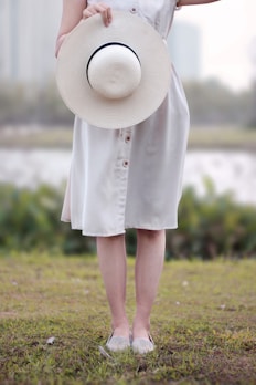A woman in a soft sand-colored dress smiling while holding a straw hat against a neutral background.