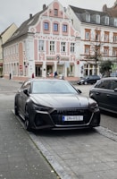 A sleek black car is parked on a cobblestone street in front of a historic European building with ornate architectural details. The building showcases pastel pink and white colors with intricate patterns. Several people can be seen walking by, and another vehicle is parked nearby.