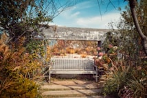 A wooden bench is placed in the center of a lush garden, surrounded by various plants and flowers in warm autumnal colors. The garden is enclosed by a stone wall, and beyond it, taller grasses and distant trees are visible. The sky is clear with a few clouds.