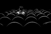 A lone individual sits facing away from the camera in a large and empty theater or cinema, surrounded by rows of empty seats. The scene is captured in black and white, creating a heightened sense of solitude.