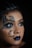 A close-up view of a person with elaborate face art. The design features colorful butterfly patterns around the eyes and a floral crescent moon on the cheek. The lips are painted a deep blue, complementing the overall artistic makeup. The background is dark, highlighting the intricate details of the face art.