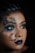 A close-up view of a person with elaborate face art. The design features colorful butterfly patterns around the eyes and a floral crescent moon on the cheek. The lips are painted a deep blue, complementing the overall artistic makeup. The background is dark, highlighting the intricate details of the face art.