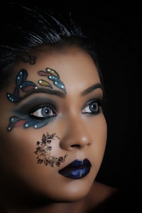 A close-up view of a person with elaborate face art. The design features colorful butterfly patterns around the eyes and a floral crescent moon on the cheek. The lips are painted a deep blue, complementing the overall artistic makeup. The background is dark, highlighting the intricate details of the face art.