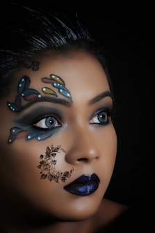 A close-up view of a person with elaborate face art. The design features colorful butterfly patterns around the eyes and a floral crescent moon on the cheek. The lips are painted a deep blue, complementing the overall artistic makeup. The background is dark, highlighting the intricate details of the face art.