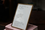 A framed piece of paper with handwritten quotes stands on top of two stacked pink books. The writing includes inspirational quotes with cited sources. The setting appears to be indoors, with some objects blurry in the background.