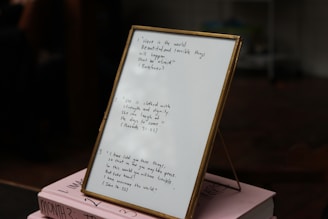 A framed piece of paper with handwritten quotes stands on top of two stacked pink books. The writing includes inspirational quotes with cited sources. The setting appears to be indoors, with some objects blurry in the background.