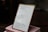 A framed piece of paper with handwritten quotes stands on top of two stacked pink books. The writing includes inspirational quotes with cited sources. The setting appears to be indoors, with some objects blurry in the background.