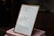 A framed piece of paper with handwritten quotes stands on top of two stacked pink books. The writing includes inspirational quotes with cited sources. The setting appears to be indoors, with some objects blurry in the background.