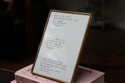 A framed piece of paper with handwritten quotes stands on top of two stacked pink books. The writing includes inspirational quotes with cited sources. The setting appears to be indoors, with some objects blurry in the background.