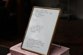 A framed piece of paper with handwritten quotes stands on top of two stacked pink books. The writing includes inspirational quotes with cited sources. The setting appears to be indoors, with some objects blurry in the background.