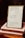 A framed quote board is resting on two stacked books with handwritten notes. The frame contains three quotes: one referencing Buethech, another citing Proverbs 31:25 about strength and dignity, and the third quoting John 16:33 about peace. The books are labeled 'MONTH 3: TRUE IDENTITY' and 'MONTH 3: IDENTITY.'