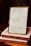 A framed quote board is resting on two stacked books with handwritten notes. The frame contains three quotes: one referencing Buethech, another citing Proverbs 31:25 about strength and dignity, and the third quoting John 16:33 about peace. The books are labeled 'MONTH 3: TRUE IDENTITY' and 'MONTH 3: IDENTITY.'