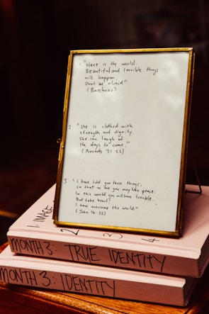 A framed quote board is resting on two stacked books with handwritten notes. The frame contains three quotes: one referencing Buethech, another citing Proverbs 31:25 about strength and dignity, and the third quoting John 16:33 about peace. The books are labeled 'MONTH 3: TRUE IDENTITY' and 'MONTH 3: IDENTITY.'