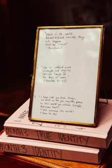 A framed quote board is resting on two stacked books with handwritten notes. The frame contains three quotes: one referencing Buethech, another citing Proverbs 31:25 about strength and dignity, and the third quoting John 16:33 about peace. The books are labeled 'MONTH 3: TRUE IDENTITY' and 'MONTH 3: IDENTITY.'