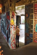 A person stands inside an urban setting filled with colorful and eclectic graffiti on concrete walls. The individual is holding a camera, capturing the surrounding artwork. The scene conveys an intriguing blend of artistic expression and exploration, with natural light streaming through an open space in the background.