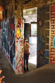 A person stands inside an urban setting filled with colorful and eclectic graffiti on concrete walls. The individual is holding a camera, capturing the surrounding artwork. The scene conveys an intriguing blend of artistic expression and exploration, with natural light streaming through an open space in the background.