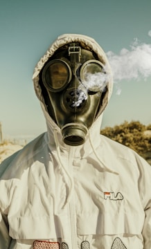 A person is wearing a gas mask with a hooded white jacket. The mask is expelling smoke or vapor, creating a mysterious or ominous effect. The scene is set outdoors with a pale blue sky in the background.