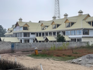 A large, multi-story building with a light-colored roof featuring numerous dormer windows, set against a cloudy sky. The building is surrounded by a low stone wall, and there is vegetation in front of the wall. A dirt path leads to the building, with a small yellow trash can visible on the side. In the background, there are tall trees and a communication tower.