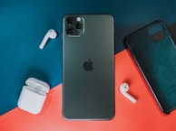 A smartphone with a triple camera setup placed on a red and teal background, accompanied by two wireless earbuds and a charging case. A phone case is positioned nearby.