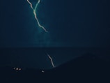 A dramatic scene of a lightning bolt cutting through a dark, stormy night sky. The jagged line of the lightning contrasts sharply with the surrounding darkness, illuminating a silhouette of a distant rooftop and faint outlines of vegetation. The atmosphere is charged and intense, capturing the raw power of a thunderstorm.