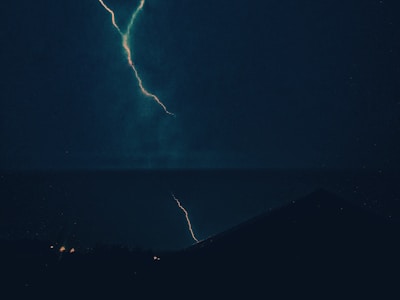 A dramatic scene of a lightning bolt cutting through a dark, stormy night sky. The jagged line of the lightning contrasts sharply with the surrounding darkness, illuminating a silhouette of a distant rooftop and faint outlines of vegetation. The atmosphere is charged and intense, capturing the raw power of a thunderstorm.