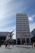 A tall, multi-story building labeled 'The Student Hotel' dominates the scene, with a modern sculpture in front and a blue sky overhead. Several people are walking around the plaza area.