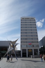 A tall, multi-story building labeled 'The Student Hotel' dominates the scene, with a modern sculpture in front and a blue sky overhead. Several people are walking around the plaza area.