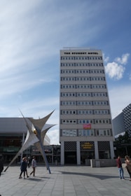 A tall, multi-story building labeled 'The Student Hotel' dominates the scene, with a modern sculpture in front and a blue sky overhead. Several people are walking around the plaza area.