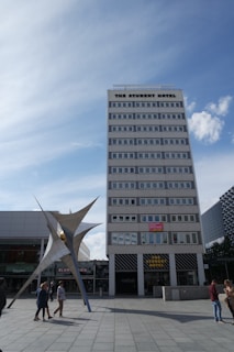 A tall, multi-story building labeled 'The Student Hotel' dominates the scene, with a modern sculpture in front and a blue sky overhead. Several people are walking around the plaza area.