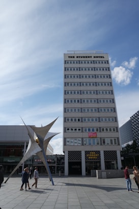 A tall, multi-story building labeled 'The Student Hotel' dominates the scene, with a modern sculpture in front and a blue sky overhead. Several people are walking around the plaza area.