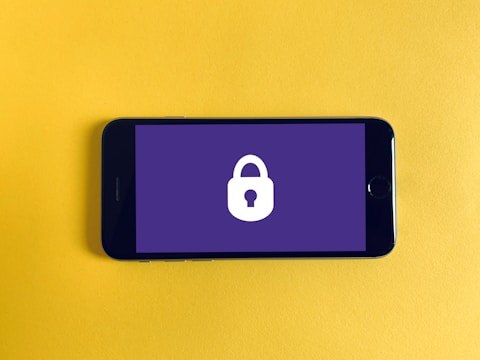 A smartphone placed on a bright yellow background displays a lock symbol on its screen, which is colored in a flat purple shade. The lock symbol suggests themes of security or privacy.