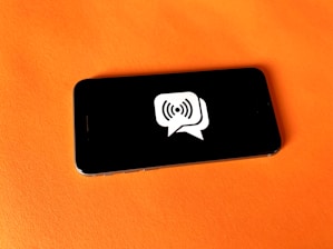 A smartphone rests on a bright orange surface. The screen displays a graphic of a white speech bubble with a wireless signal icon inside.