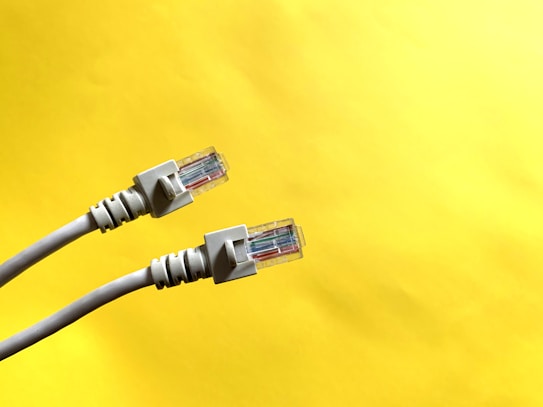 Two Ethernet cables with transparent connectors are positioned against a bright yellow background. The cables are light gray, with details visible in the wiring within the connectors.