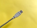 A USB cable with a gray connector and metallic casing on the cord, positioned diagonally against a bright yellow background.