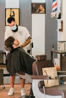A barber wearing a mask is trimming a client's beard in a vintage-style barbershop. The client is seated in a reclining chair covered with a black cape. The barber has tattoos on his arms and is focused on his work. In the background, framed photos adorn the walls, and a classic barber pole is visible. A cardboard box is placed on a nearby chair.