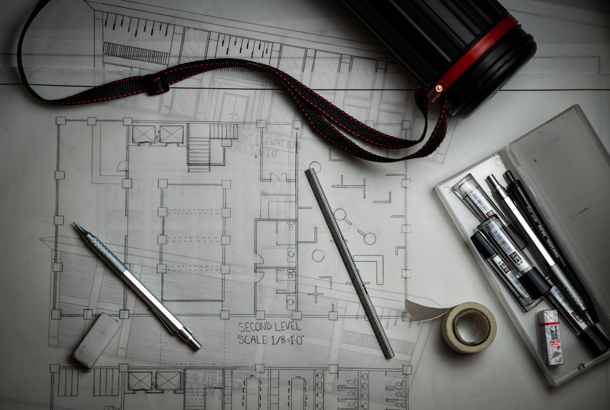 Architectural blueprints laid out on a desk with drafting pencils, a metal ruler and a drawing tube