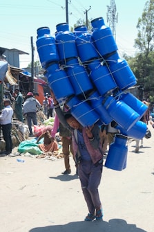 A person is carrying a large load of blue plastic containers tied together on their back while walking through a busy market area. Surrounding the individual are various people and market stalls filled with goods. The scene is bustling with activity, and there are buildings and trees visible in the background.