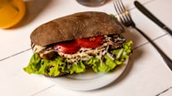 A sandwich with dark bread is filled with fresh green lettuce, slices of red tomato, shredded cheese, and what appears to be grilled chicken. The sandwich is placed on a white plate with a knife and fork nearby on a wooden table.