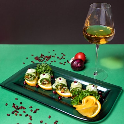 A stylish arrangement of gourmet appetizers with rolled cucumber slices filled with a vegetable mixture on a dark green rectangular plate. Garnished with orange slices, parsley, and pomegranate seeds. A glass of white wine is positioned to the right, and whole vegetables, including a tomato, onion, and garlic, are scattered around.