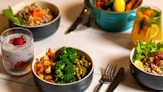 A table is set with several dishes containing a variety of healthy foods. There are bowls filled with colorful salads, vegetables like broccoli and peas, grains topped with vegetables, and glasses of infused water garnished with strawberry slices. A fork and knife are placed on the table, suggesting a meal ready to be enjoyed.